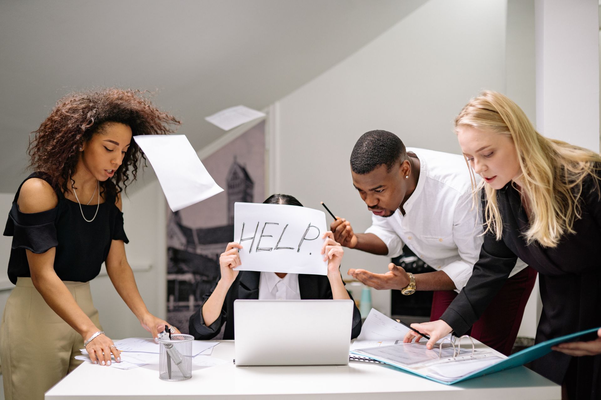 Stressed office team overwhelmed at a desk, one person holding a paper that says Help